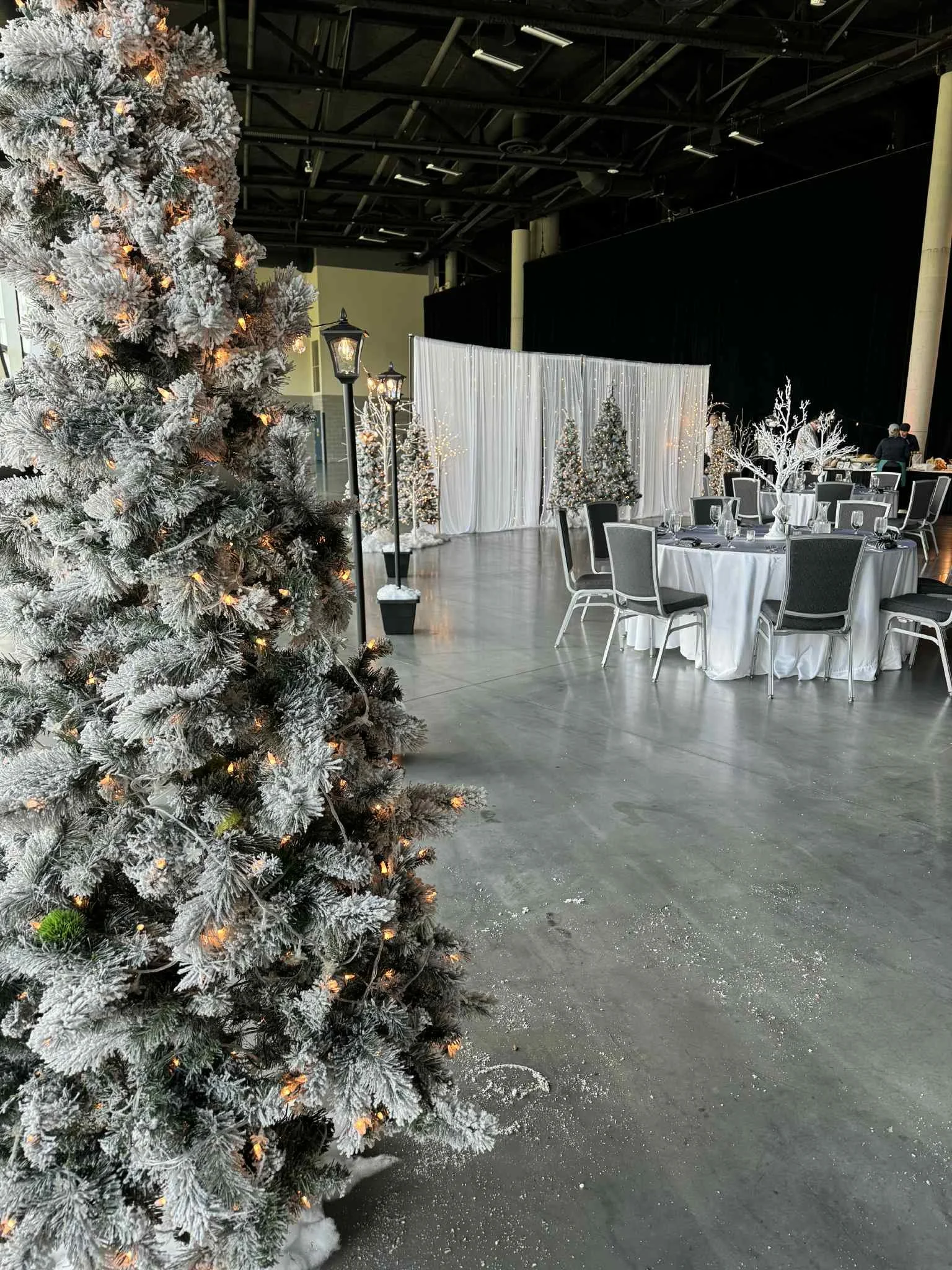 An indoor holiday event setup with decorated Christmas trees, white draped backdrop, round tables with white tablecloths and black chairs, and snow-like decorations on the floor.