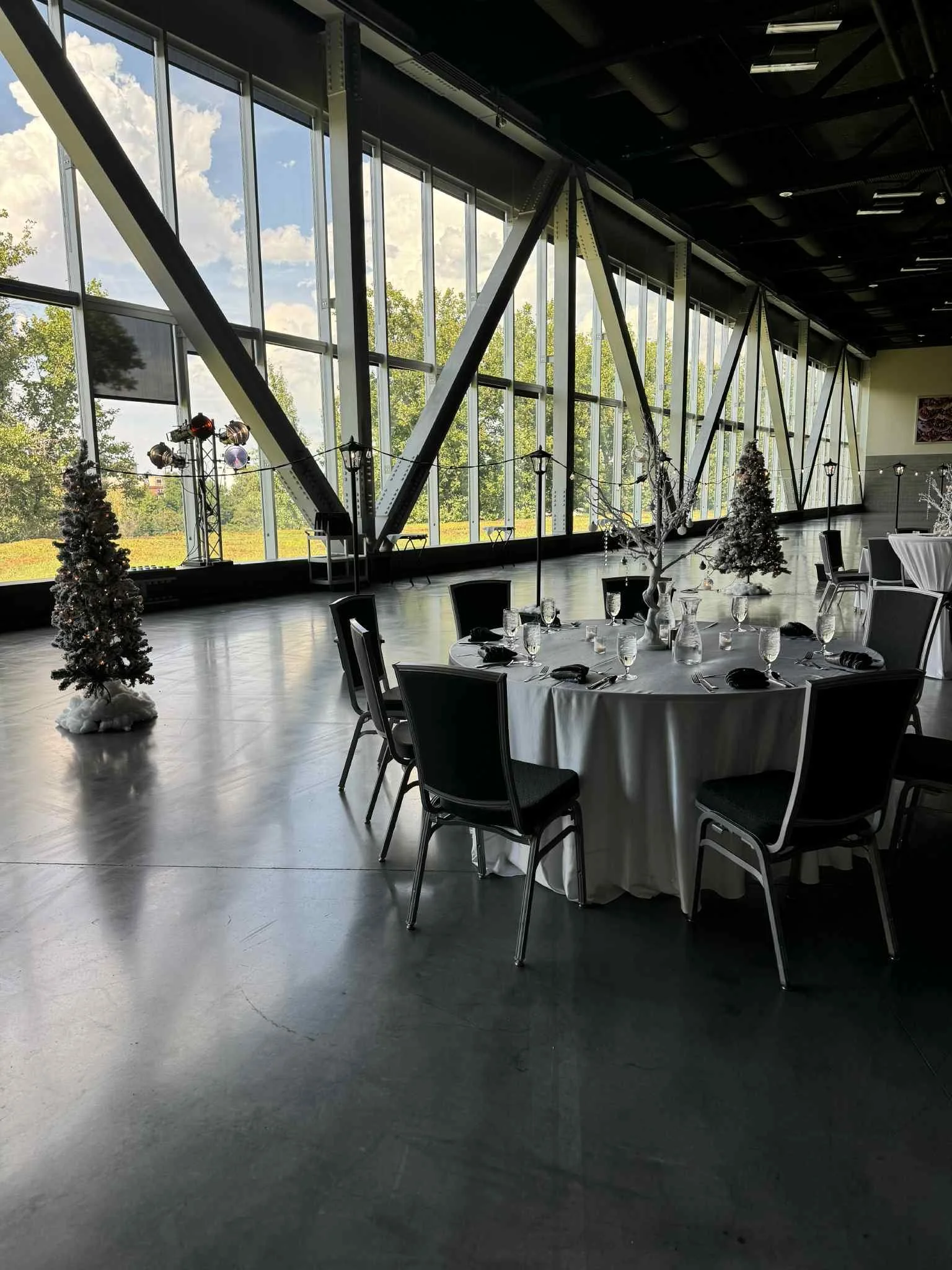 Indoor event space decorated for a holiday celebration with a round table set with glassware, black napkins, and a winter-themed centerpiece, large windows showing trees and a partly cloudy sky outside, Christmas trees with white decorations, and a d