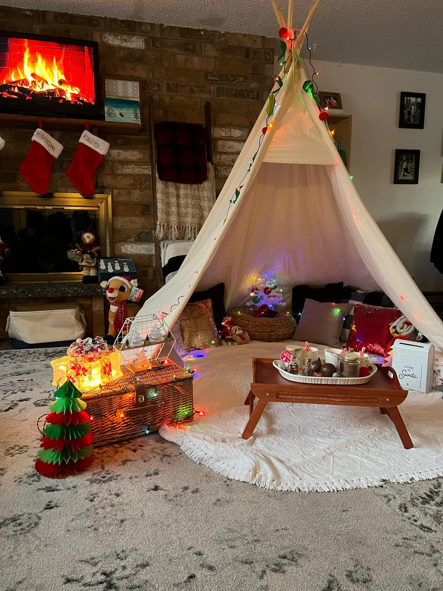 Christmas-themed living room with a beige teepee decorated with colorful string lights, small Christmas tree, and ornaments inside. A tray with holiday treats and a small festive basket sit on a wooden table in front. In the background, a brick firep
