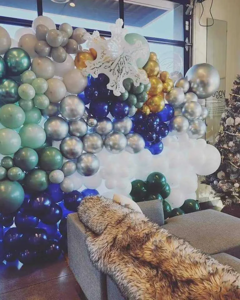 A large balloon display with gold, silver, white, navy blue, and green balloons arranged in a cluster behind a gray couch. A white decorative snowflake cutout and a Christmas tree are also visible in the scene, indicating a holiday decoration setup.