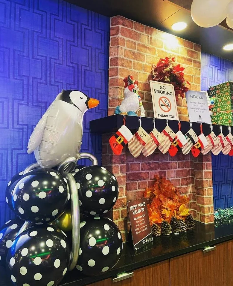 Decorative Christmas display with a festive penguin balloon, a ceramic turkey, holiday stockings, and ornaments on a mantle with holiday signs and decorations, including a "No Smoking" sign and autumn leaves with pinecones.