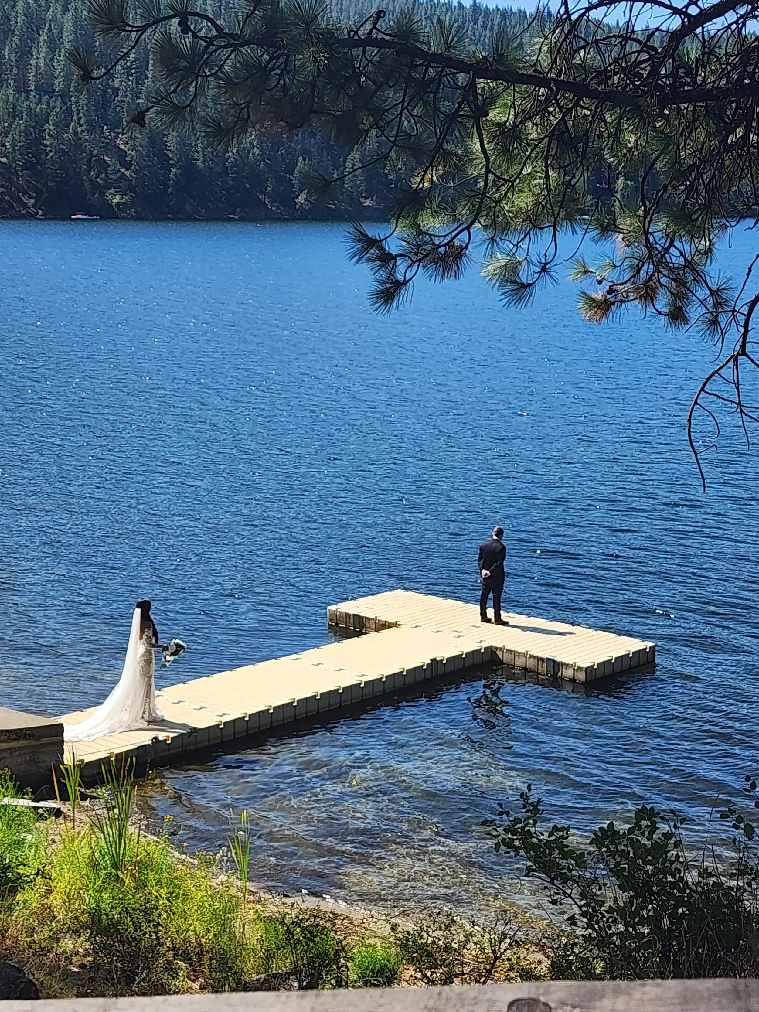 A bride in a white wedding gown holding flowers and a groom in black standing on a wooden dock by a lake surrounded by trees.