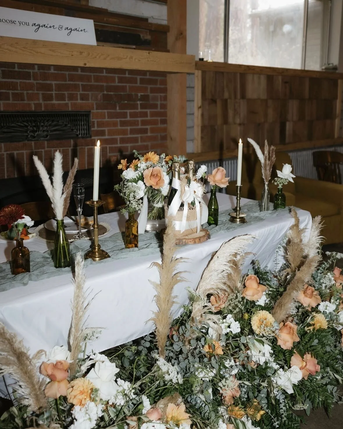 Decorated table with floral arrangements, candles, and bottles, set for a special event or celebration in a rustic-style venue.