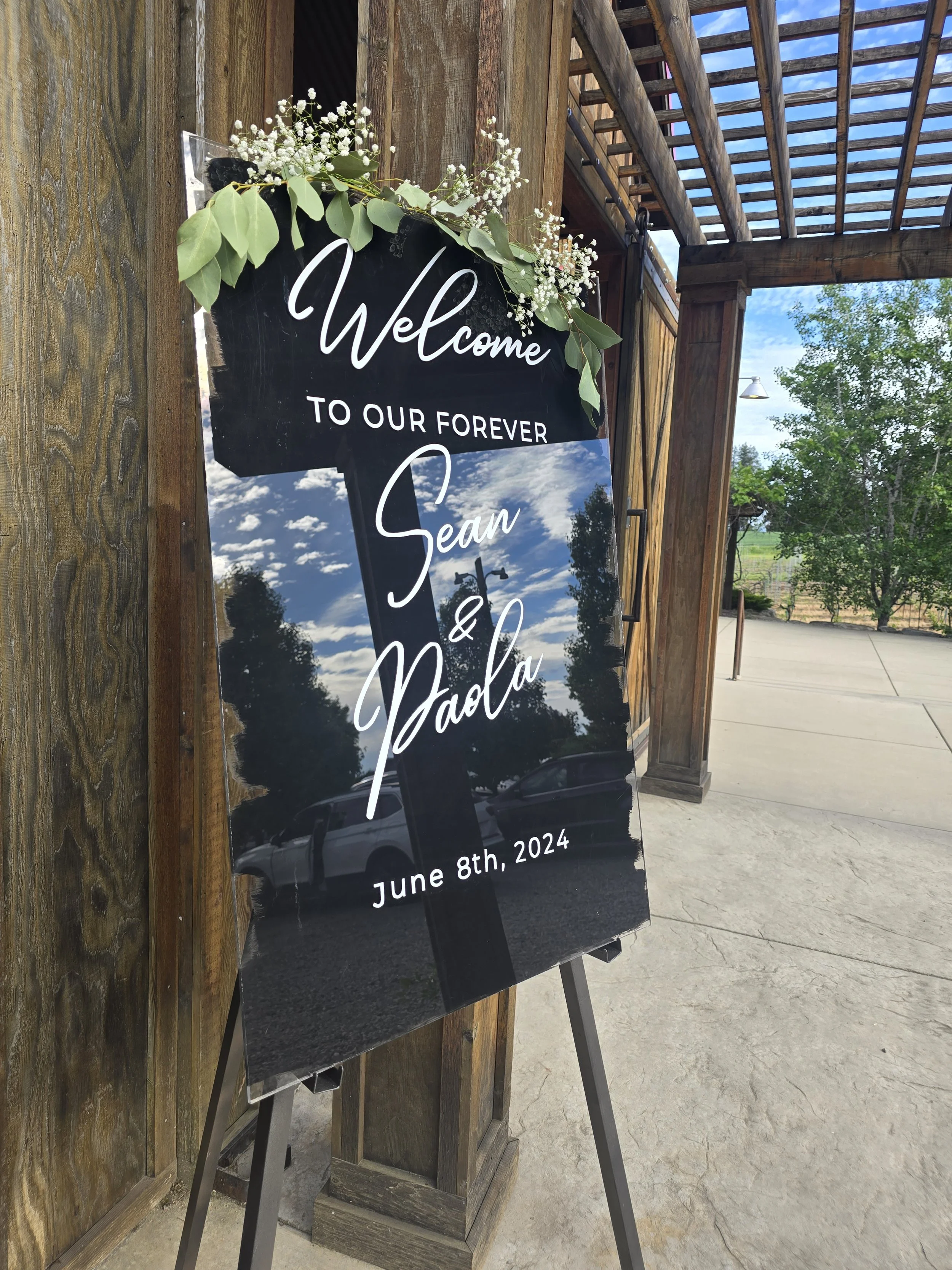 Wedding welcome sign with black background and white text, decorated with green leaves and small white flowers, displaying the names Sean and Paola and the date June 8th, 2024.