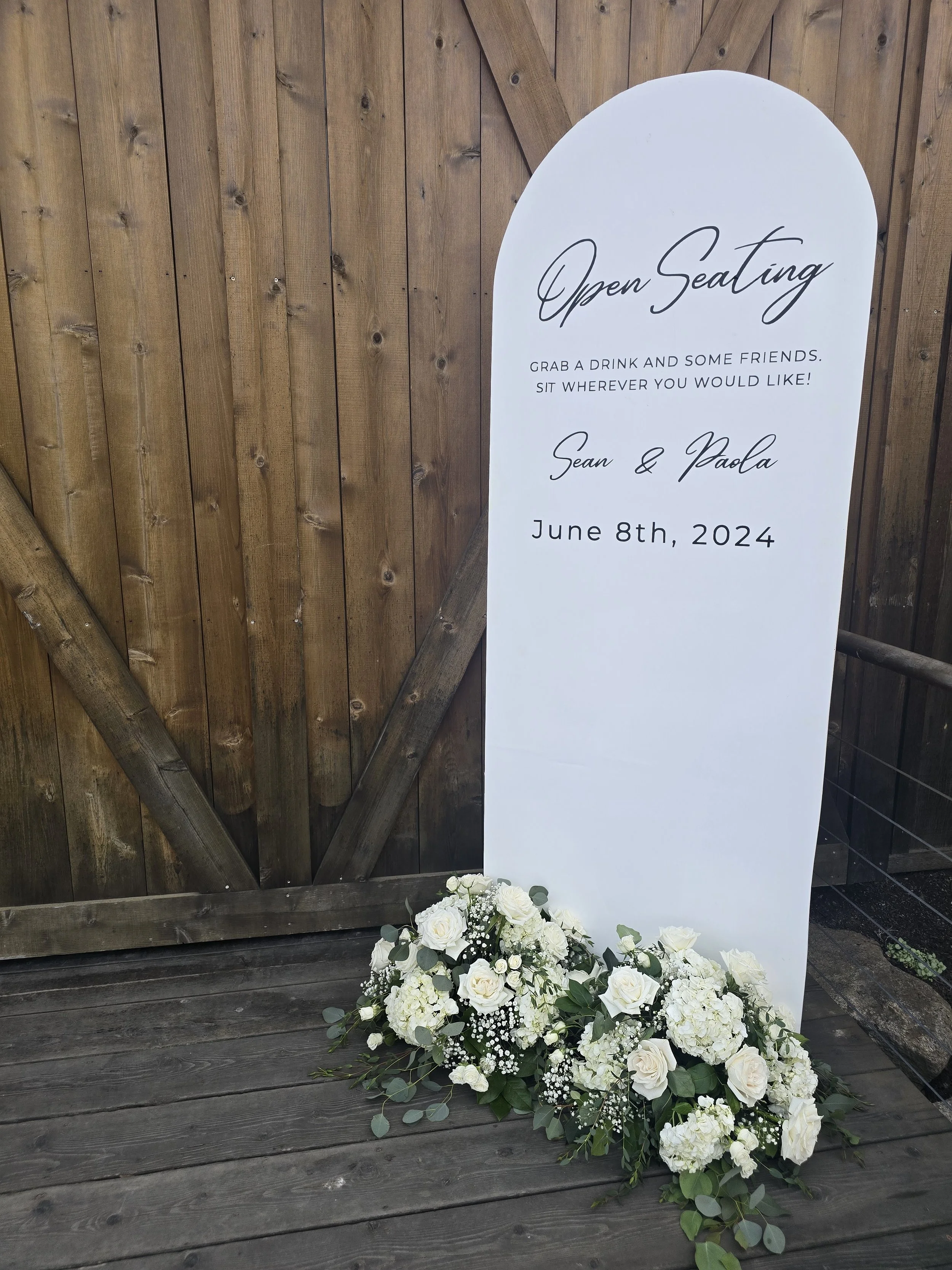 White wedding sign with black cursive and print text, decorated with white flowers and greenery at the base, leaning against a wooden fence and deck.
