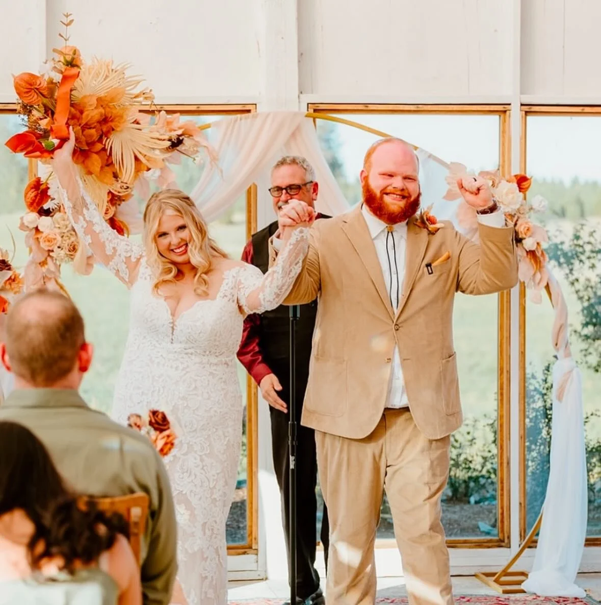 Wedding ceremony with bride and groom holding hands, smiling, and celebrating indoors with floral decorations and guests in the foreground.
