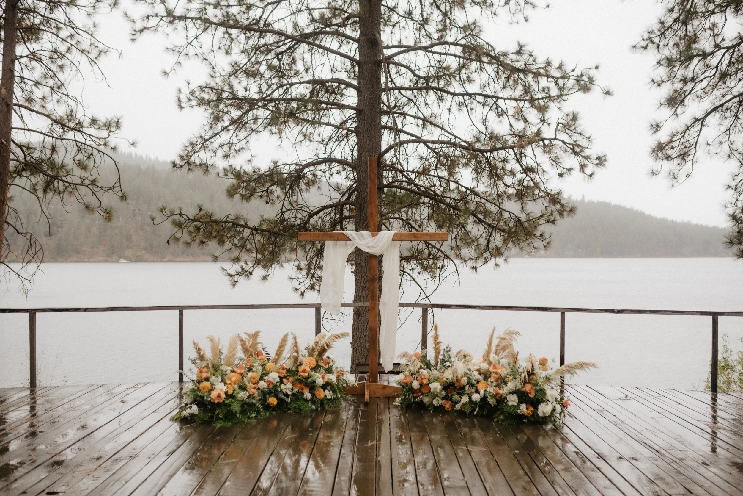 Outdoor wedding altar with a wooden cross draped with white fabric, surrounded by floral arrangements on a wooden deck by a lake, framed by pine trees.