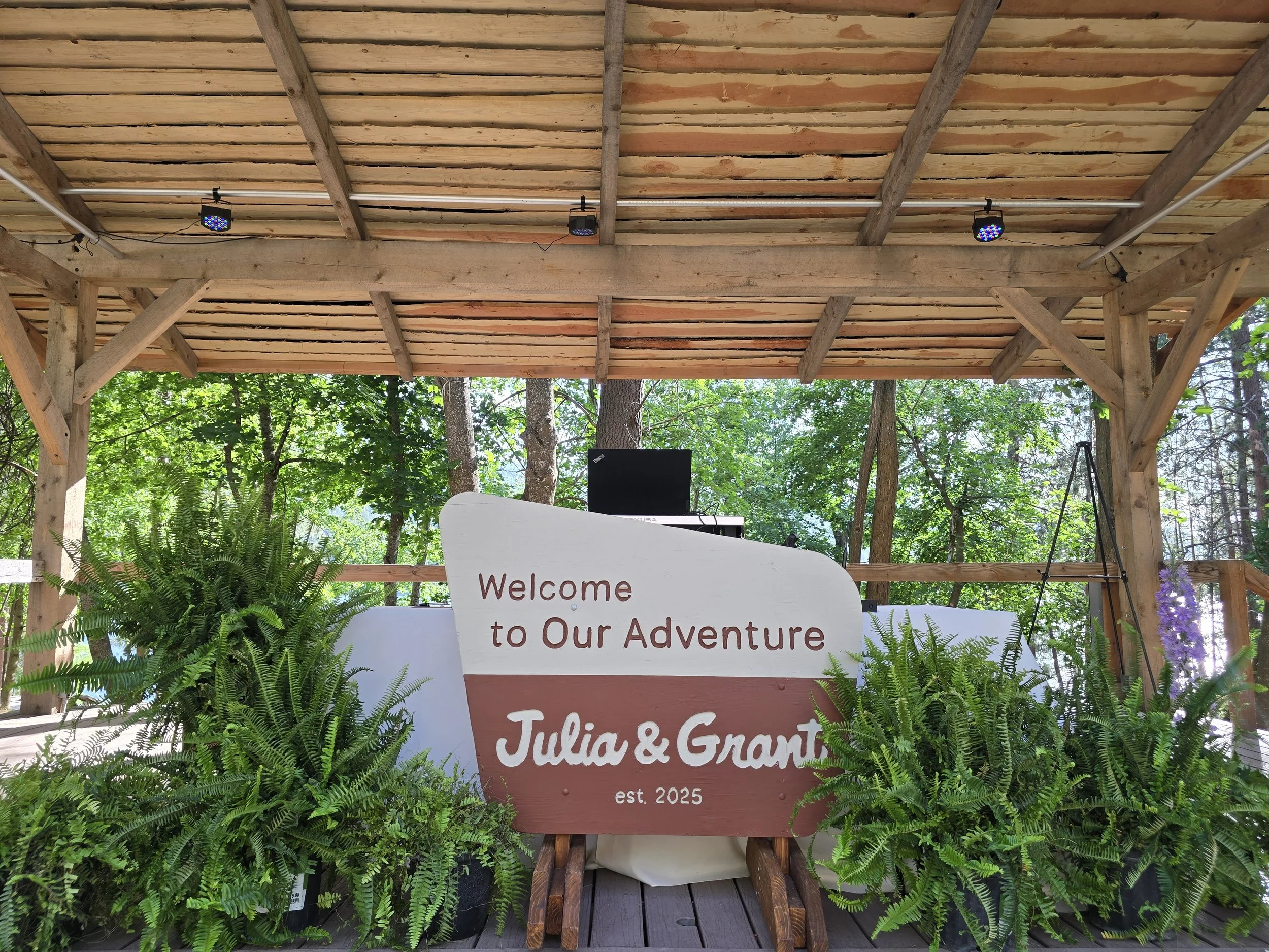 Wedding or event reception area with a wooden stage, sign with a welcome message for Julia & Grant, surrounded by green ferns and trees in the background under a wooden roof.