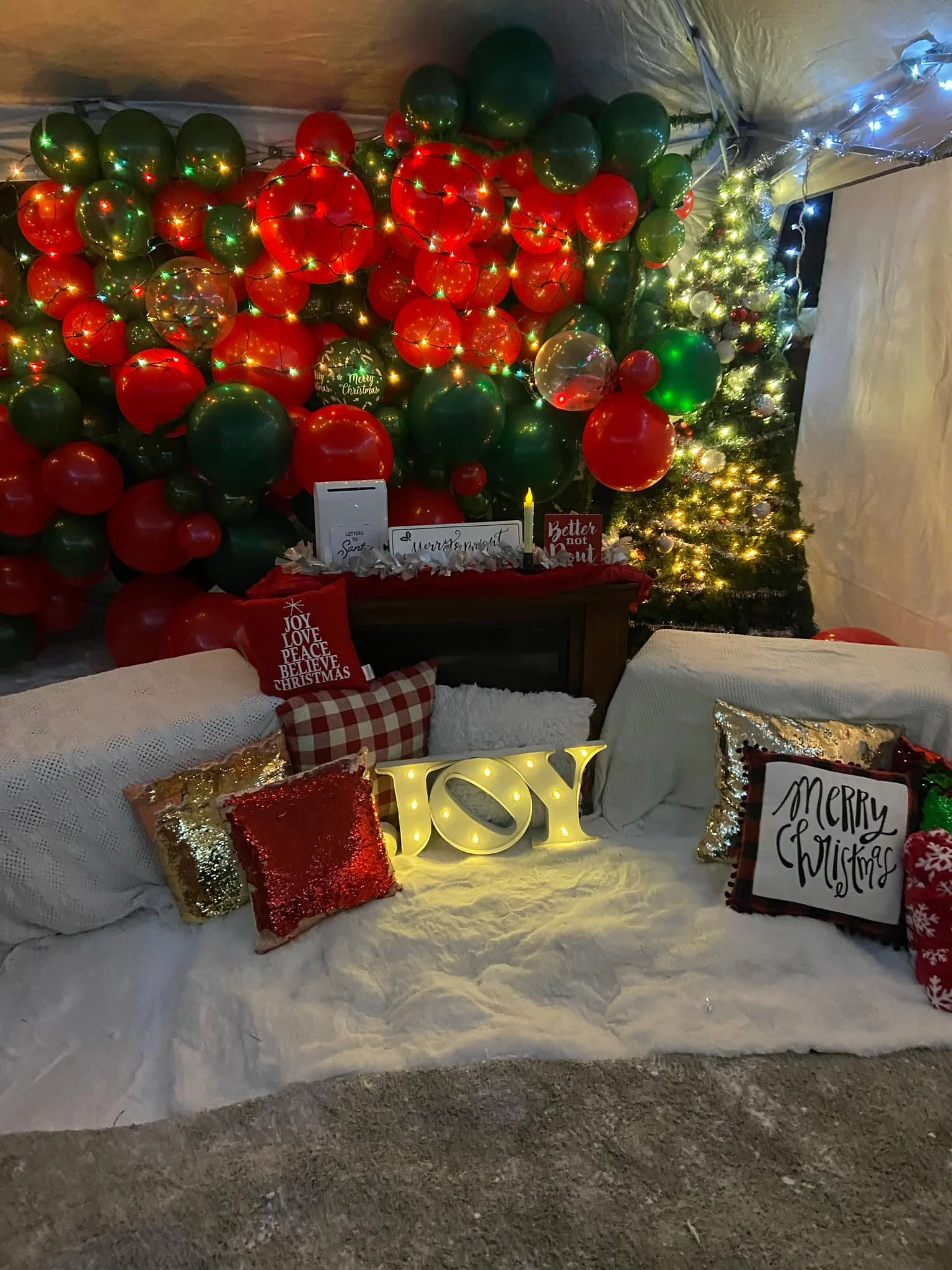 A decorated Christmas scene with a large arrangement of red and green balloons and a small Christmas tree with lights. In front, there are holiday pillows, a white blanket, and a decorative 