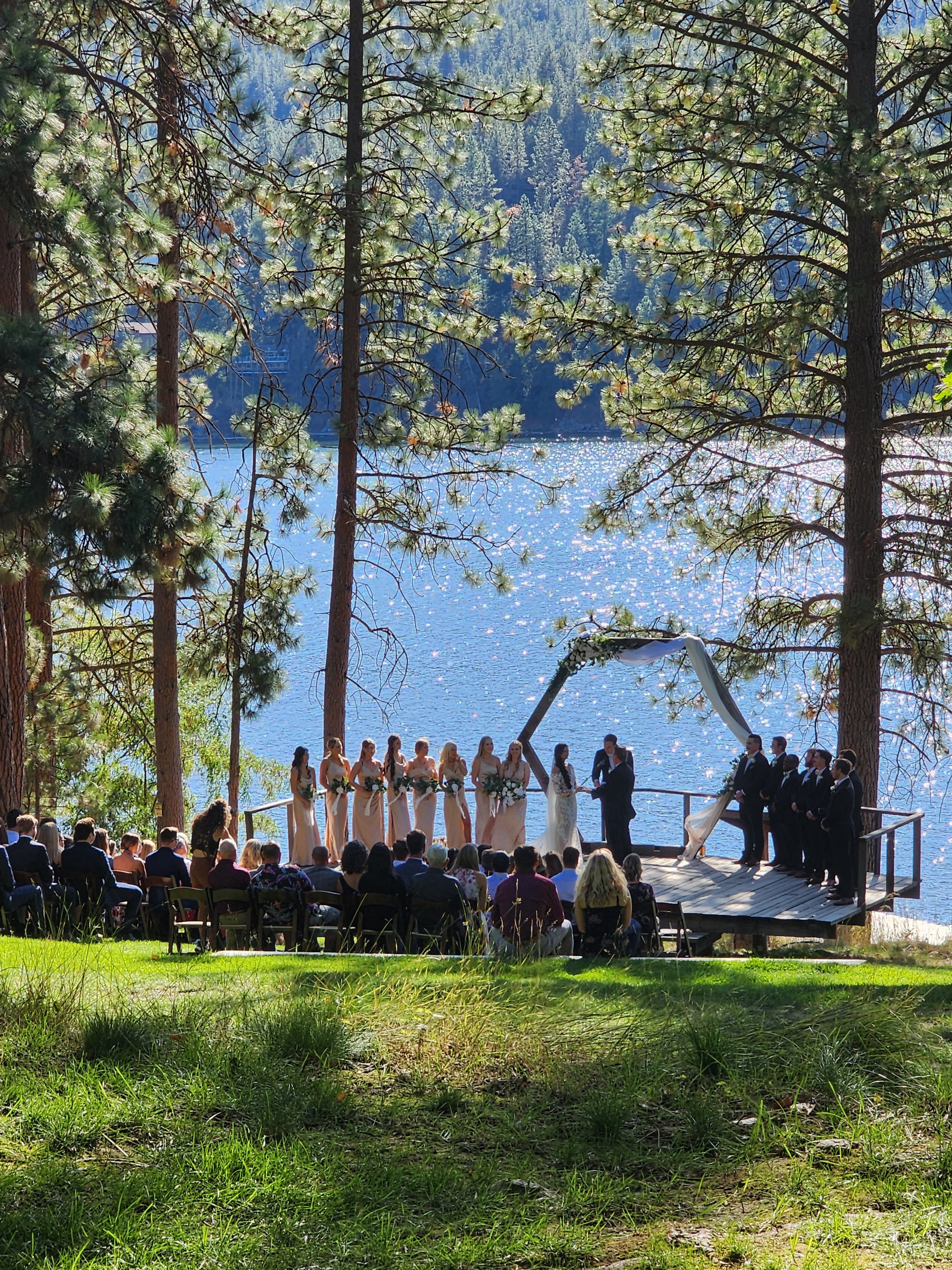 Outdoor wedding ceremony by a lake with trees surrounding the area. The bride and groom are exchanging vows under a decorated arch on a wooden platform, with bridesmaids and groomsmen standing beside them. Guests are seated watching the ceremony.