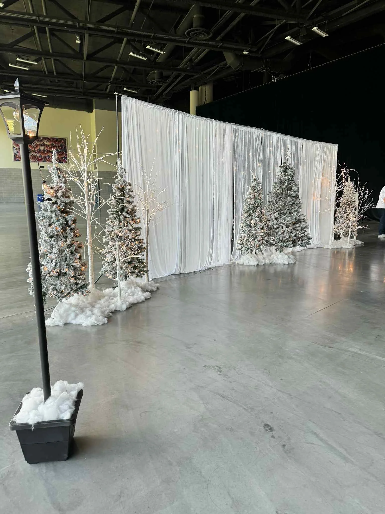 Indoor winter scene decoration with artificial snow on the ground, snow-covered Christmas trees, leafless white trees, and white fabric drapes as a backdrop, with a street lamp on the left side.