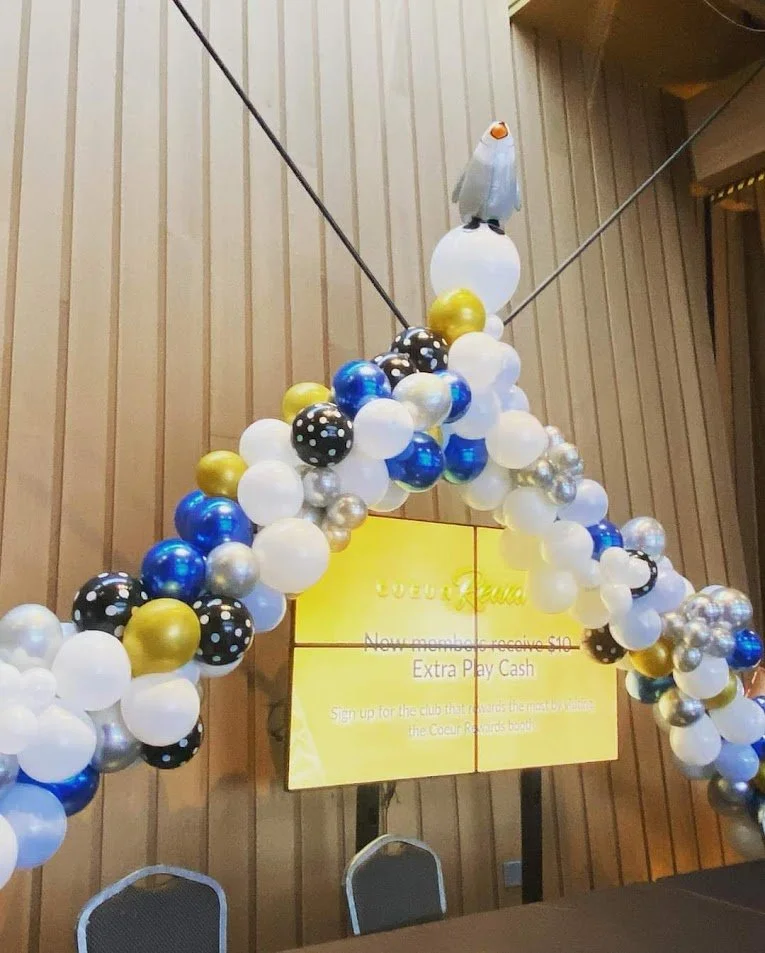 Balloon arch with a snowman figure on top, decorated with blue, white, black, gold, and silver balloons, next to a yellow screen showing text, in a wood-paneled room.