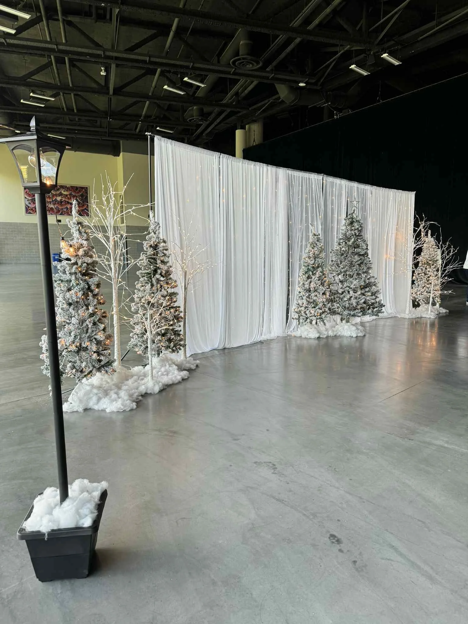 Christmas-themed backdrop with snow-covered trees, white drapery, and string lights inside a large indoor space.