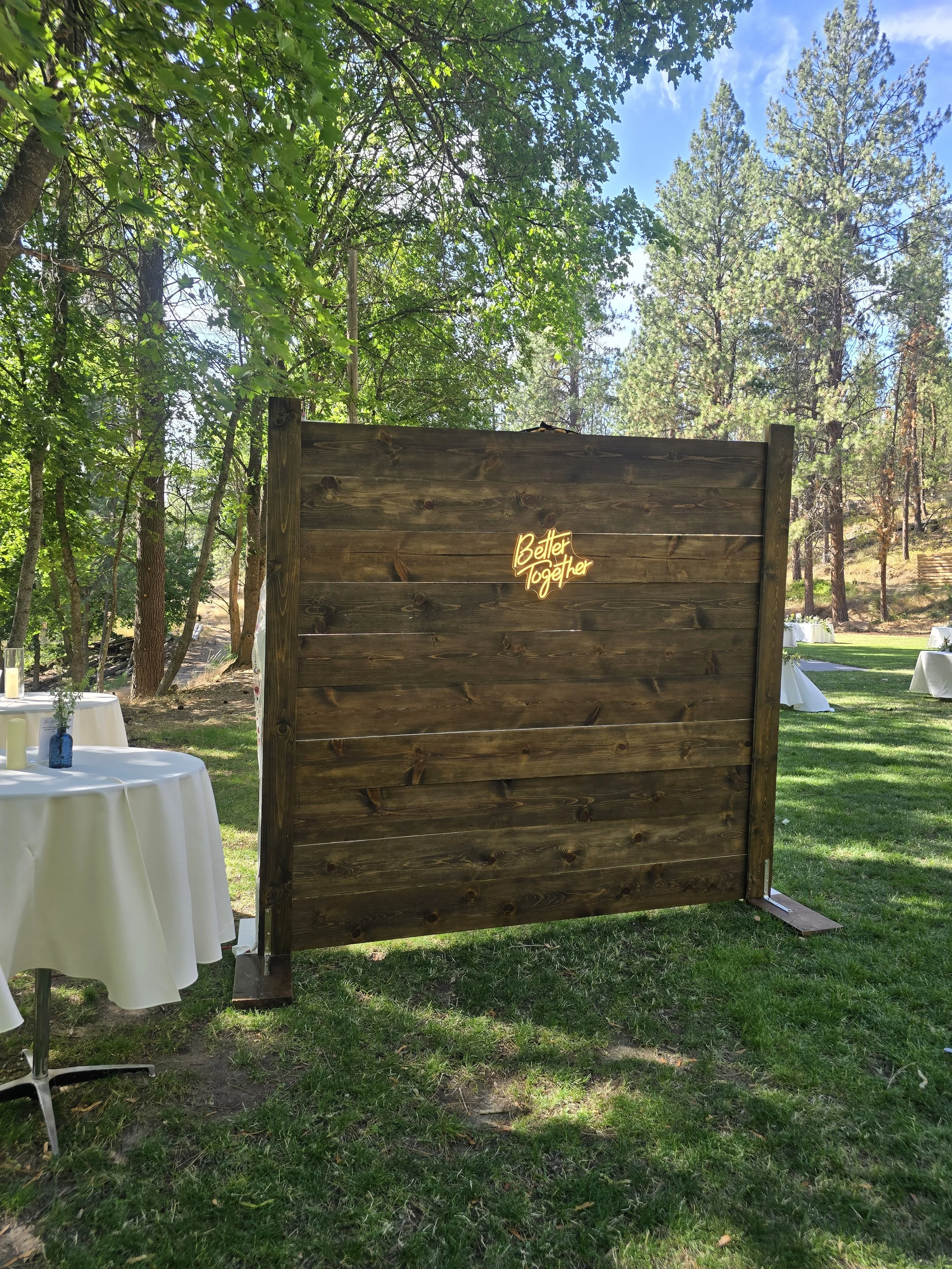 Wooden outdoor wall with a neon sign that says 'Better Together' surrounded by trees and white tables at a daytime event.