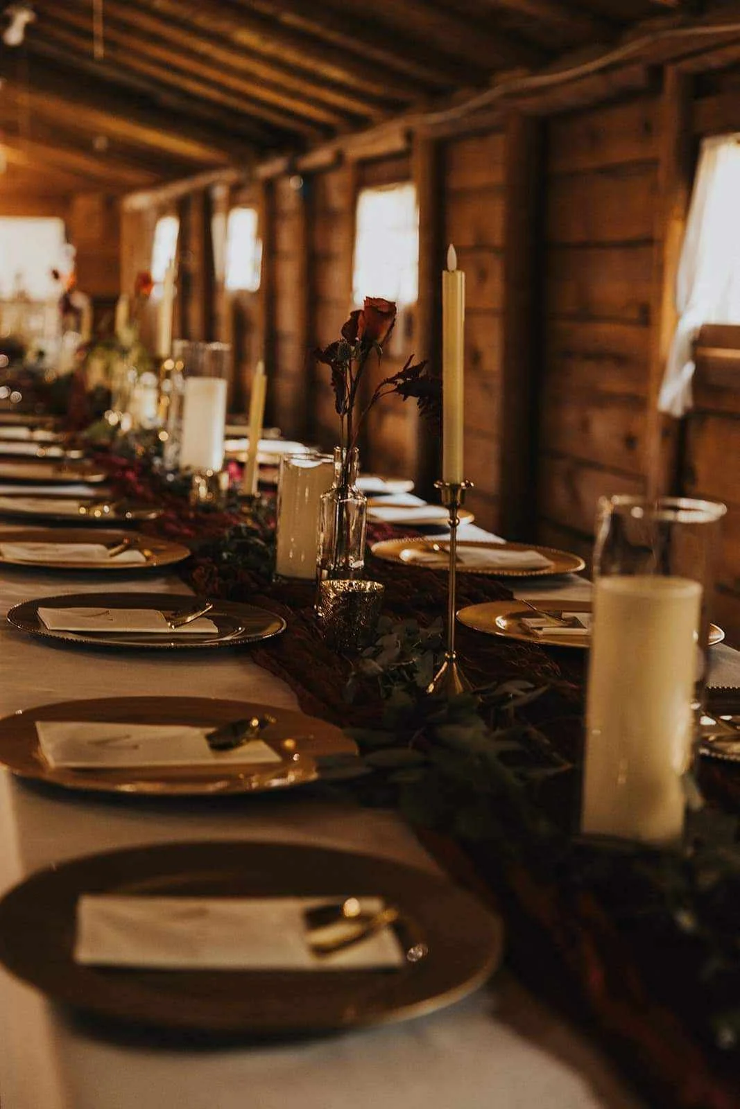 A rustic dinner table set with candles, roses, and fine china, inside a wooden cabin with warm lighting.