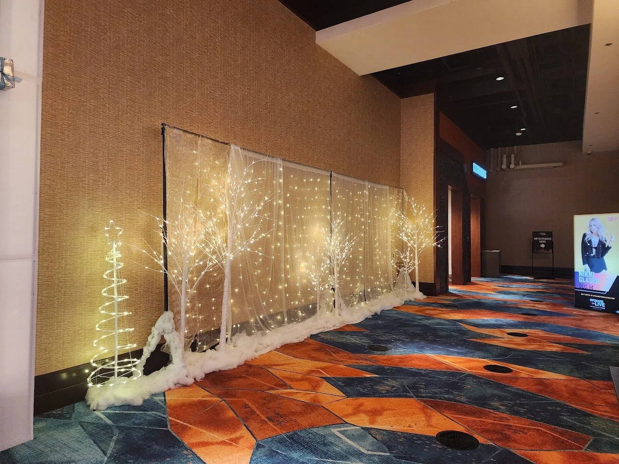 Decorative indoor display with illuminated white trees and string lights, cotton-like snow at the base, set against a beige wall and carpeted floor.