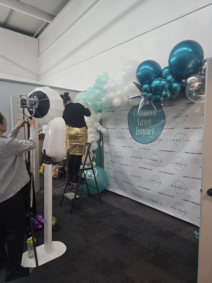 People setting up a photo backdrop with balloons for a themed event or photoshoot, featuring a large banner with the text "Connected Grow Impact".