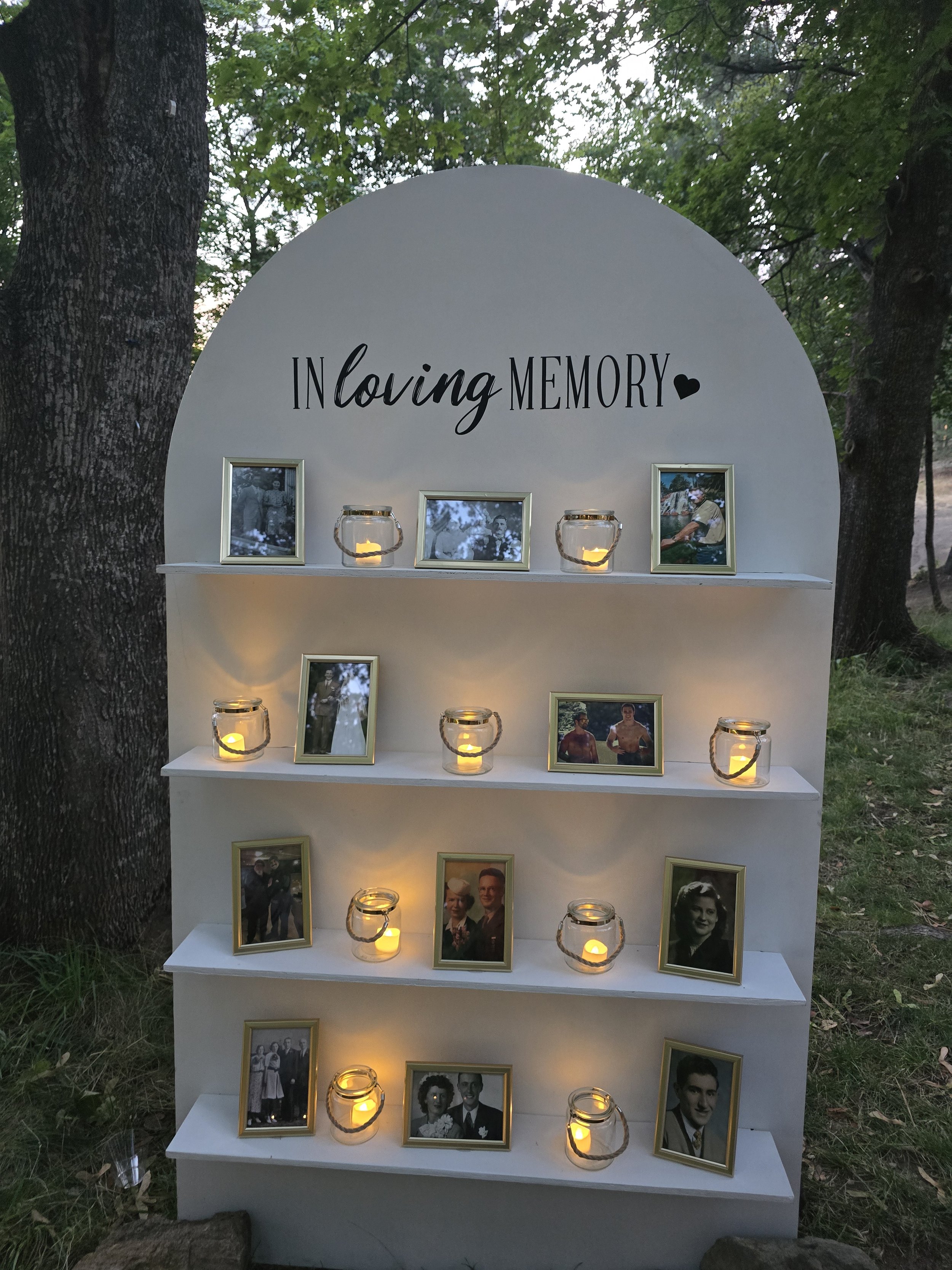 A memorial display with framed photographs, candles, and the words 'In loving MEMORY' on a white curved background, set outdoors among trees.