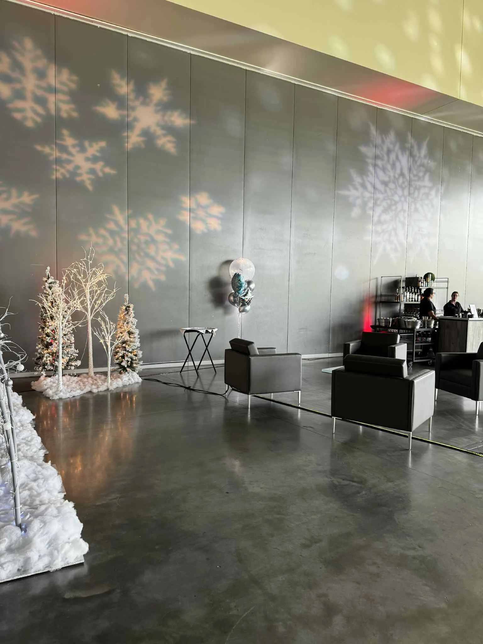 A modern indoor space with holiday decorations including white trees with snow, balloons, and snowflake light projections on a gray wall. There are black chairs, a small table, and a bar area with staff in the background.