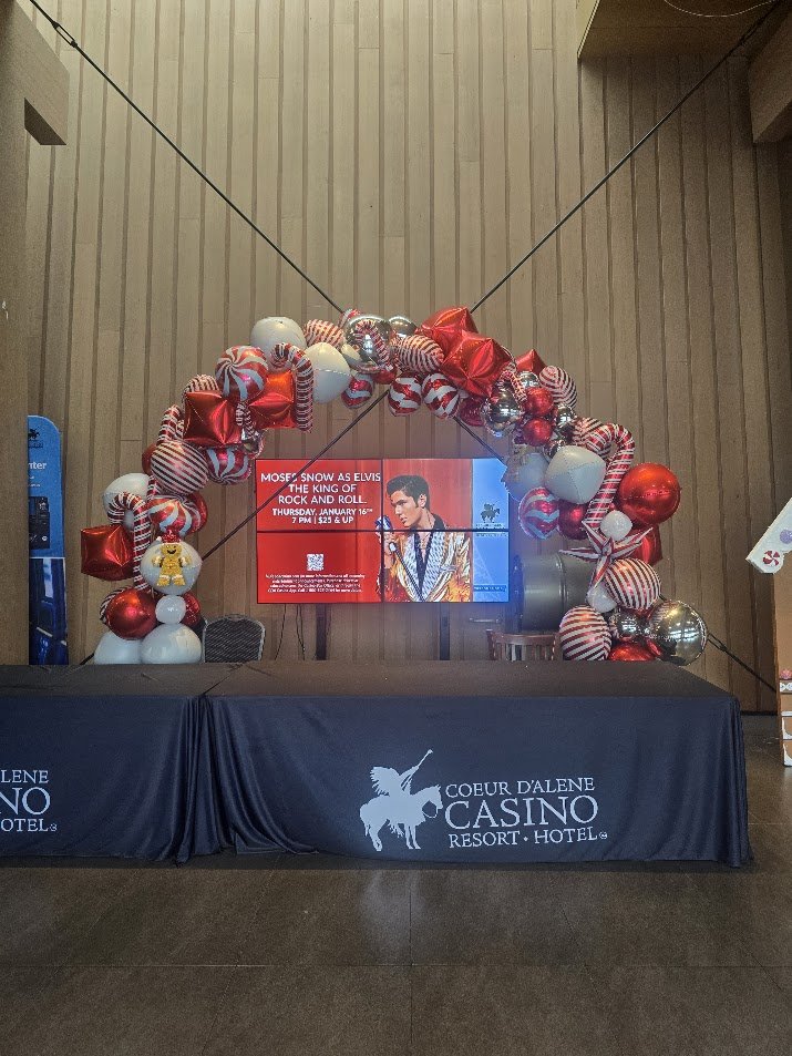 A decorated arch of Christmas ornaments, balloons, and ribbons surrounds a digital sign in a hotel lobby, with a table featuring the Coeur D'Alene Casino Resort Hotel logo in the foreground.