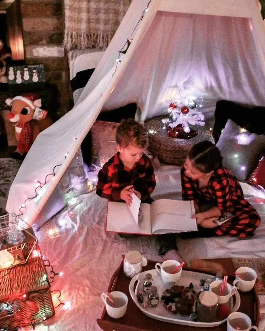 Two children sitting inside a cozy indoor tent decorated with string lights, looking at a book together. The tent has a small white Christmas tree with red ornaments and a faux fur rug. A tray with cups and snacks is in the foreground, and a plush re