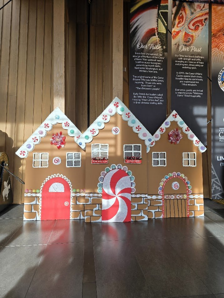 Decorative gingerbread house display with colorful icing, peppermint candy patterns, and windows, set against a wooden background with informational signs.