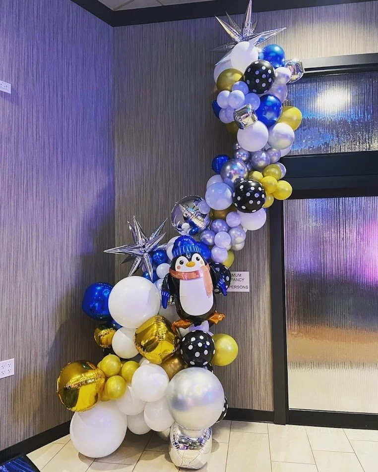 Colorful balloon arrangement with balloons in black, white, gold, blue, and silver, involving star-shaped and round balloons. Includes a penguin-shaped balloon in a blue winter hat, as part of a festive or celebratory display.