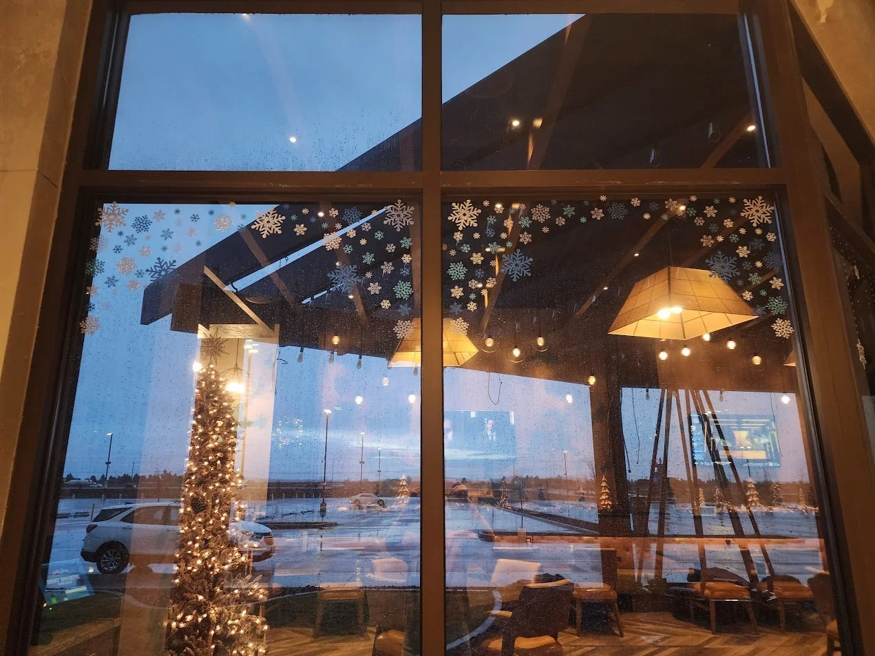 Interior view of a cafe or hotel lobby decorated for Christmas, with snowflake decals on the window and a decorated Christmas tree outside. There are warm lighting fixtures and large windows showing a rainy parking lot with cars outside.