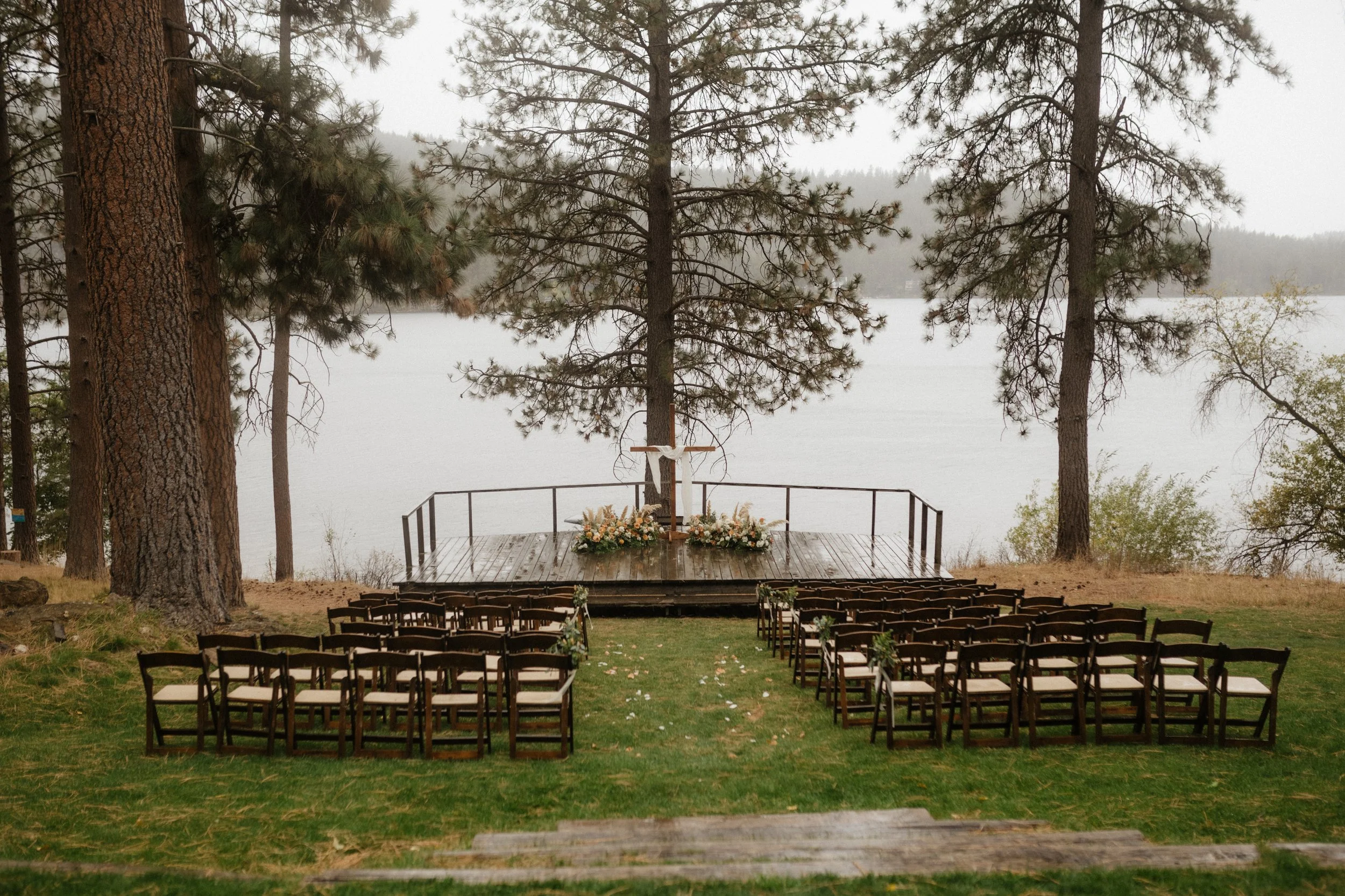 Outdoor lakeside wedding ceremony setup with chairs facing an altar decorated with flowers and a cross, surrounded by tall pine trees.
