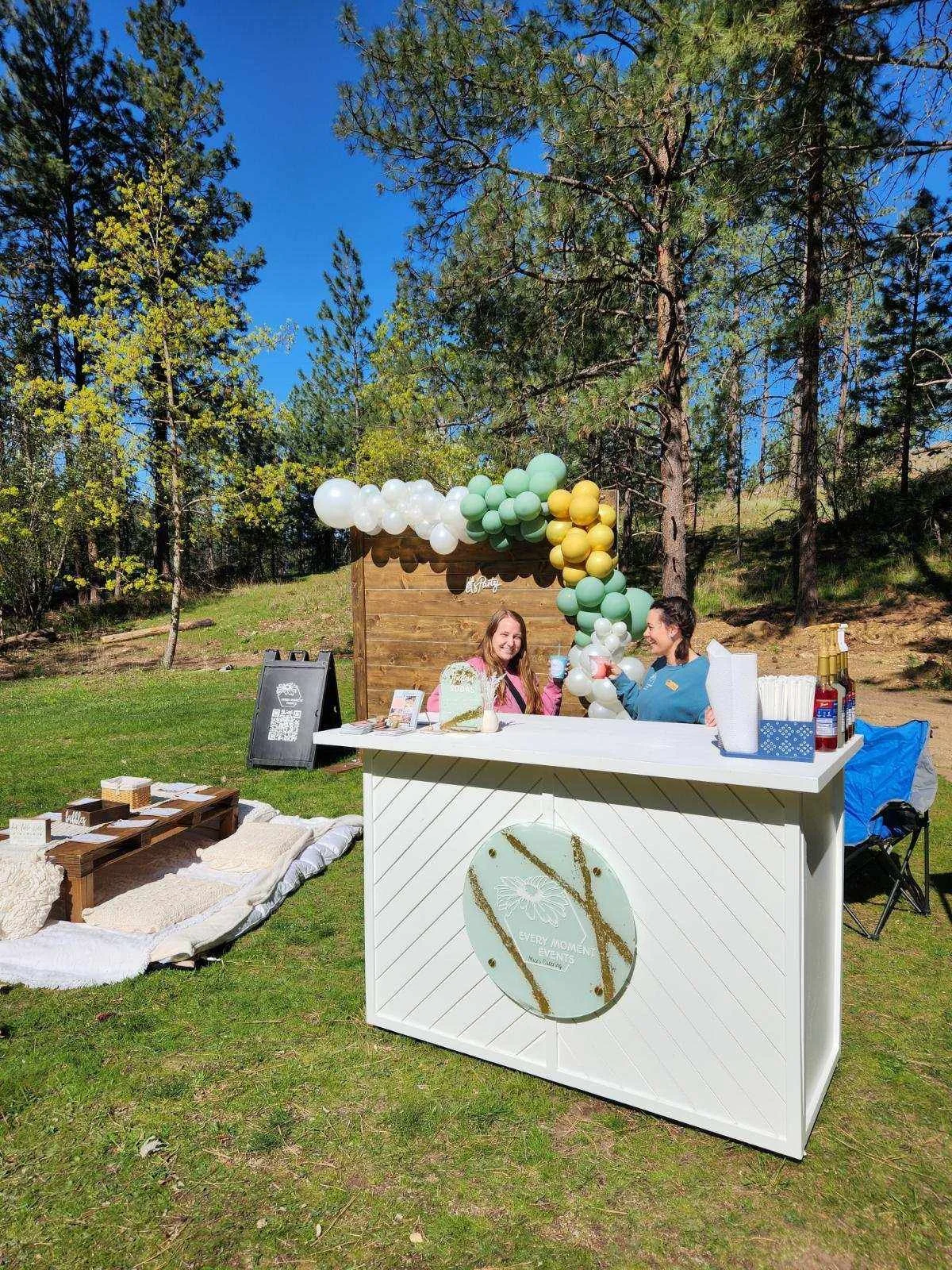 Two women standing behind a white beverage stand with balloons and outdoor trees in background, outdoors during daytime.