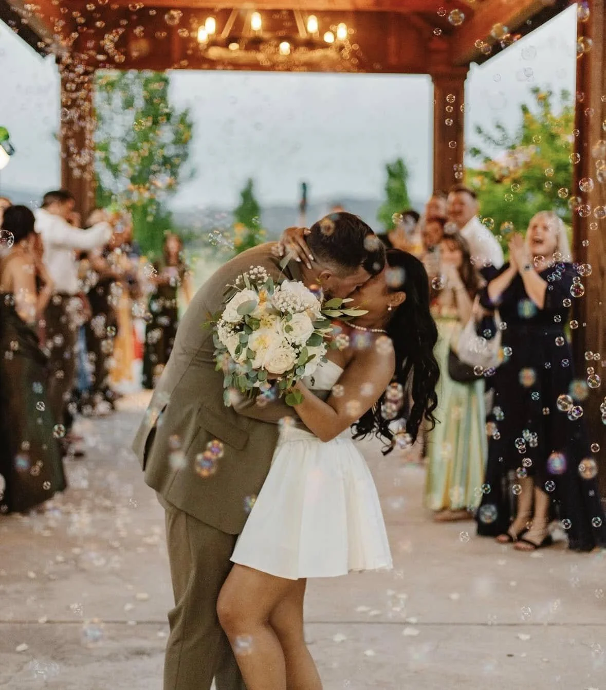 A wedding ceremony with a bride and groom kissing under a wooden pavilion decorated with lights, surrounded by guest celebrating and blowing bubbles, with outdoor greenery in the background.