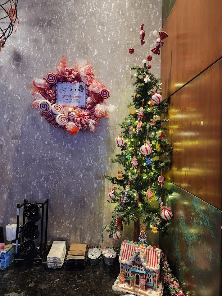 Decorated small Christmas tree with red, pink, and white candy-themed ornaments, a gingerbread house at the base, and a peppermint wreath with a sign that reads "Santa Claus The Sweet Hope of Candy Cane Lane" on a wall