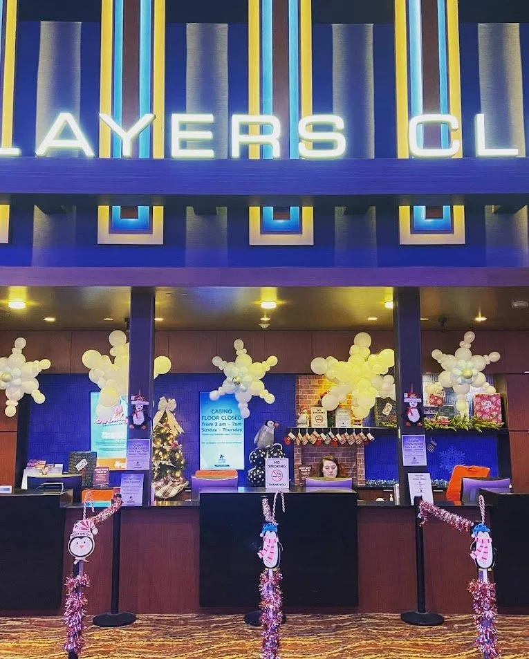Decorated hotel or casino lobby with Christmas decorations, including balloons and tinsel, behind a reception desk where a woman is working.