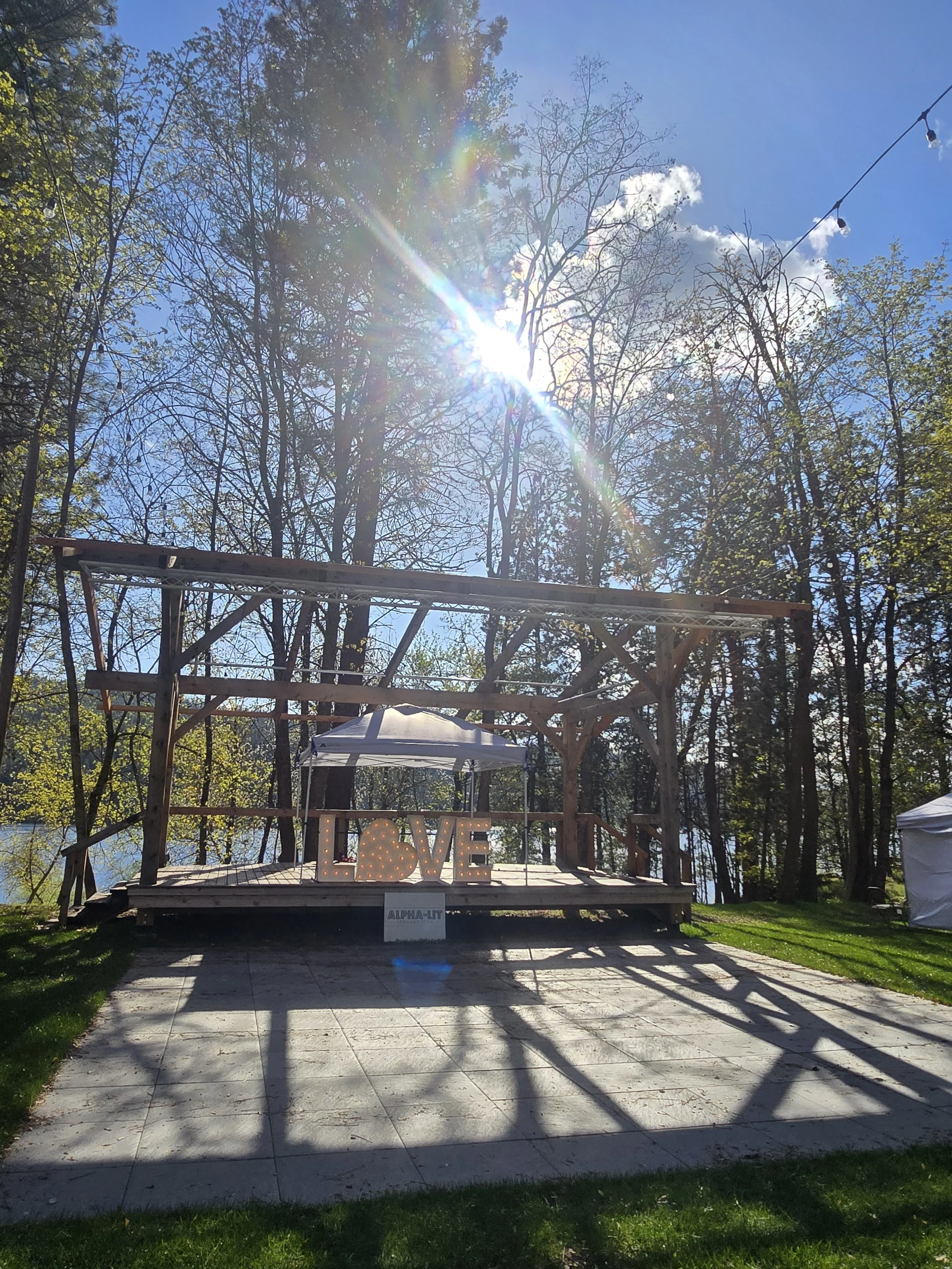 An outdoor wooden stage with a large illuminated love sign, a white canopy, and a scenic lake background under a blue sky with sunlight and trees.