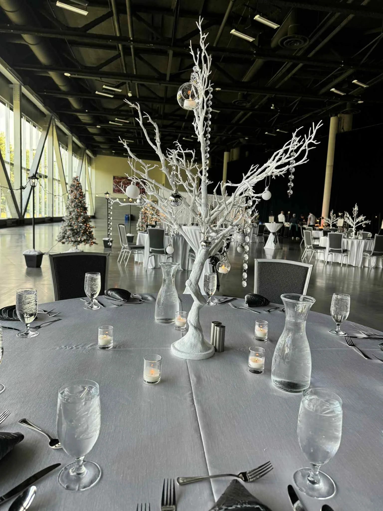Elegant banquet table setup with a white centerpiece, candles, glassware, and silverware in a large decorated hall, likely for a winter or holiday event.
