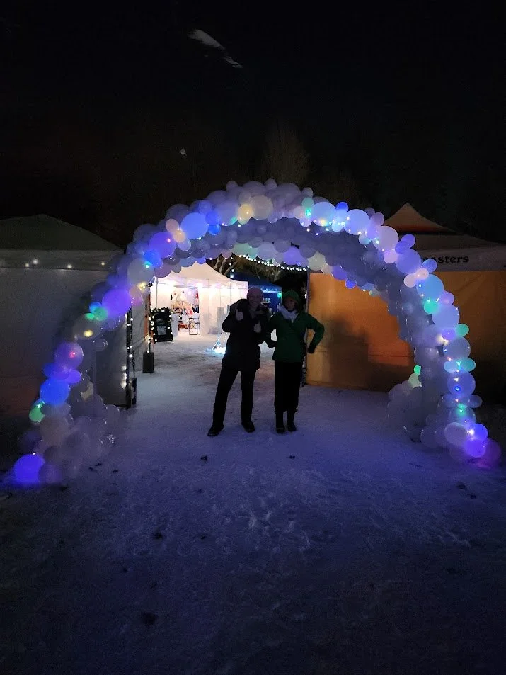 Nighttime scene with a balloon arch made of white and illuminated balloons, with two people standing underneath it. Tents and string lights are visible in the background.