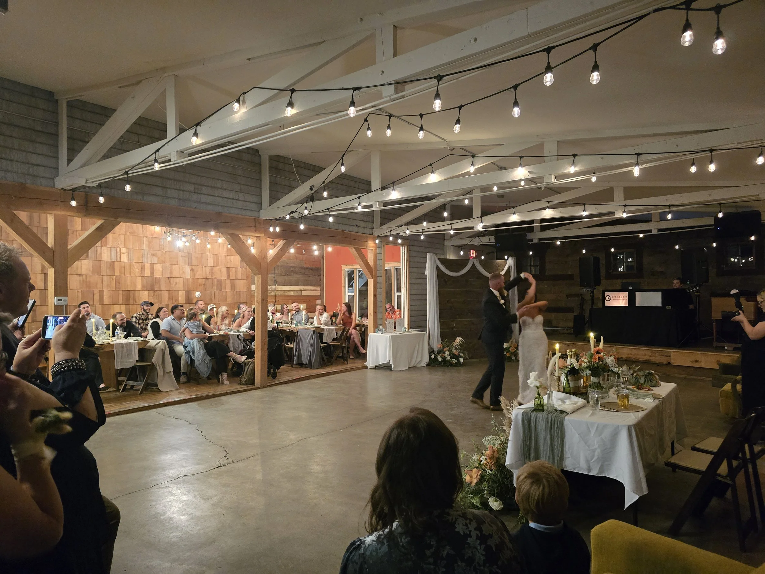 A wedding reception inside a rustic venue with string lights overhead, where a newlywed couple is dancing, and guests are seated at tables in the background.