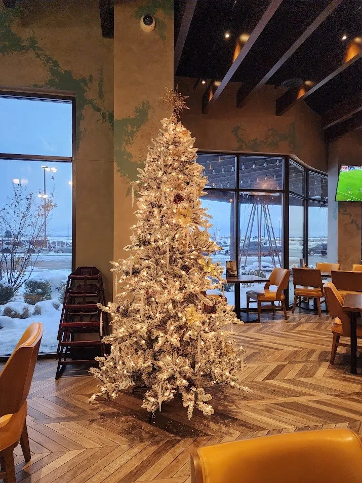 Decorated white artificial Christmas tree inside a restaurant with wooden floors and large windows showing a snowy outdoor scene.