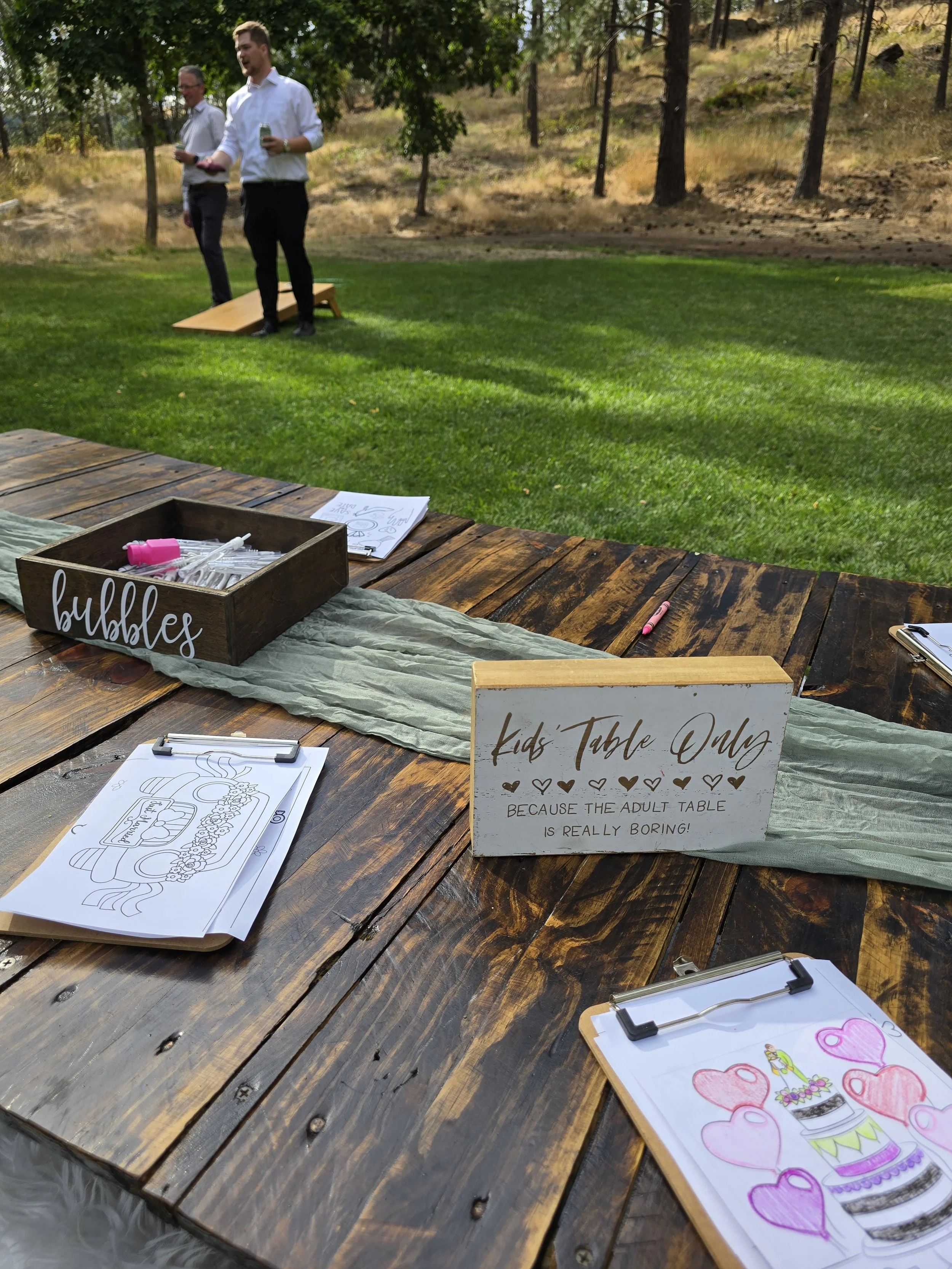 Outdoor kids' table area at a wedding, with coloring pages featuring wedding cakes and hearts, and signs indicating the kids' table and adult table, with two men standing in the background on a grassy area surrounded by trees.