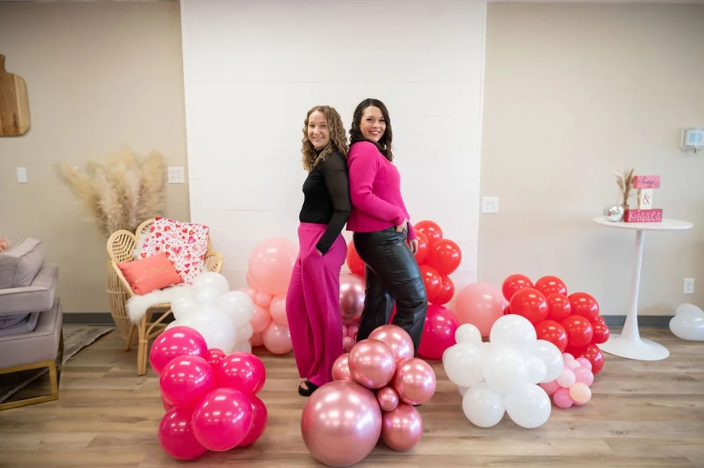 Two women stand back-to-back, smiling, amid a colorful balloon display of pink, white, and metallic balloons in a decorated indoor setting.