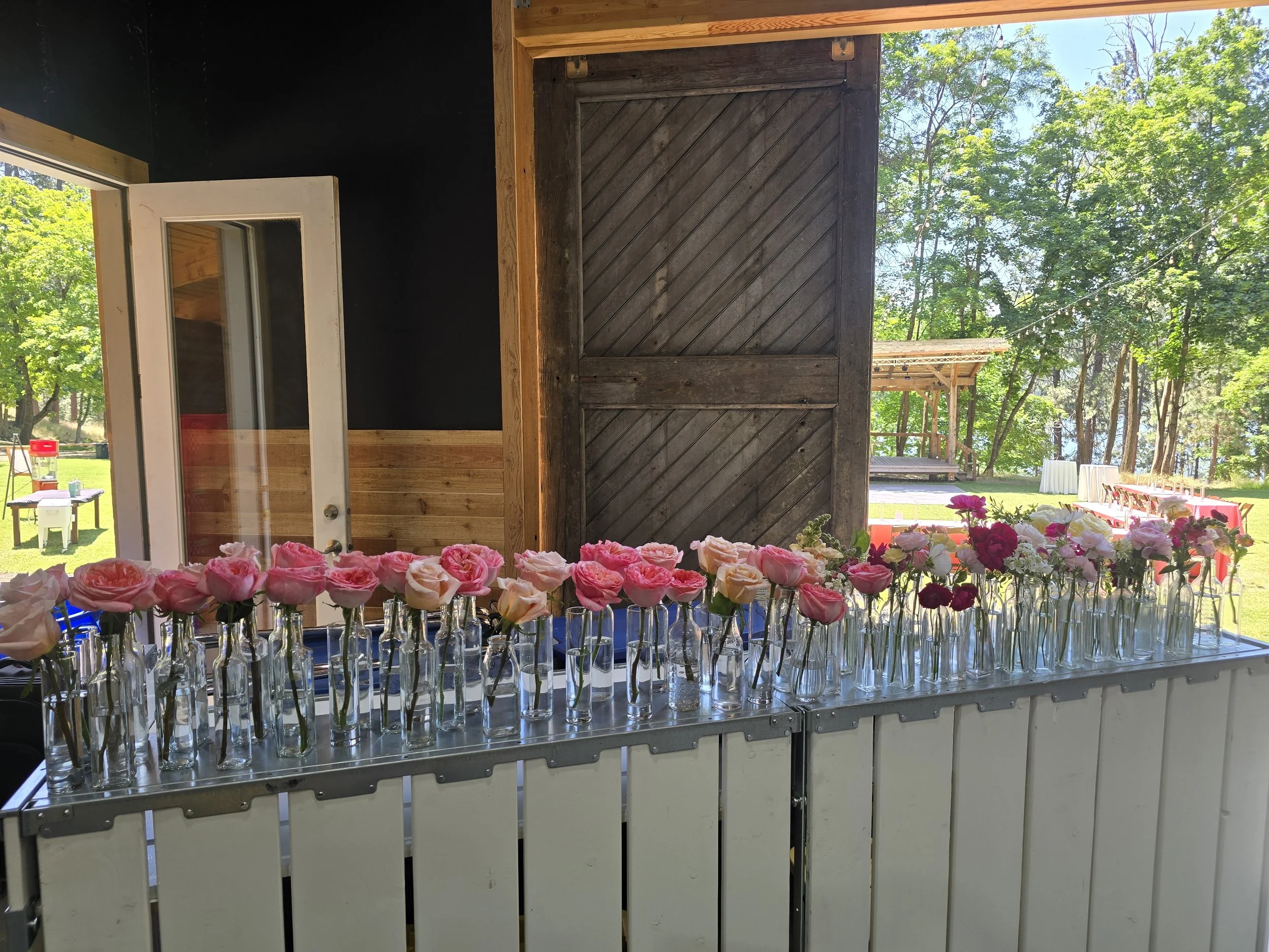 Table with small glass bottles filled with pink, white, and red roses, arranged in a line inside a rustic barn or shed with open double doors and outdoor seating area visible through the opening.