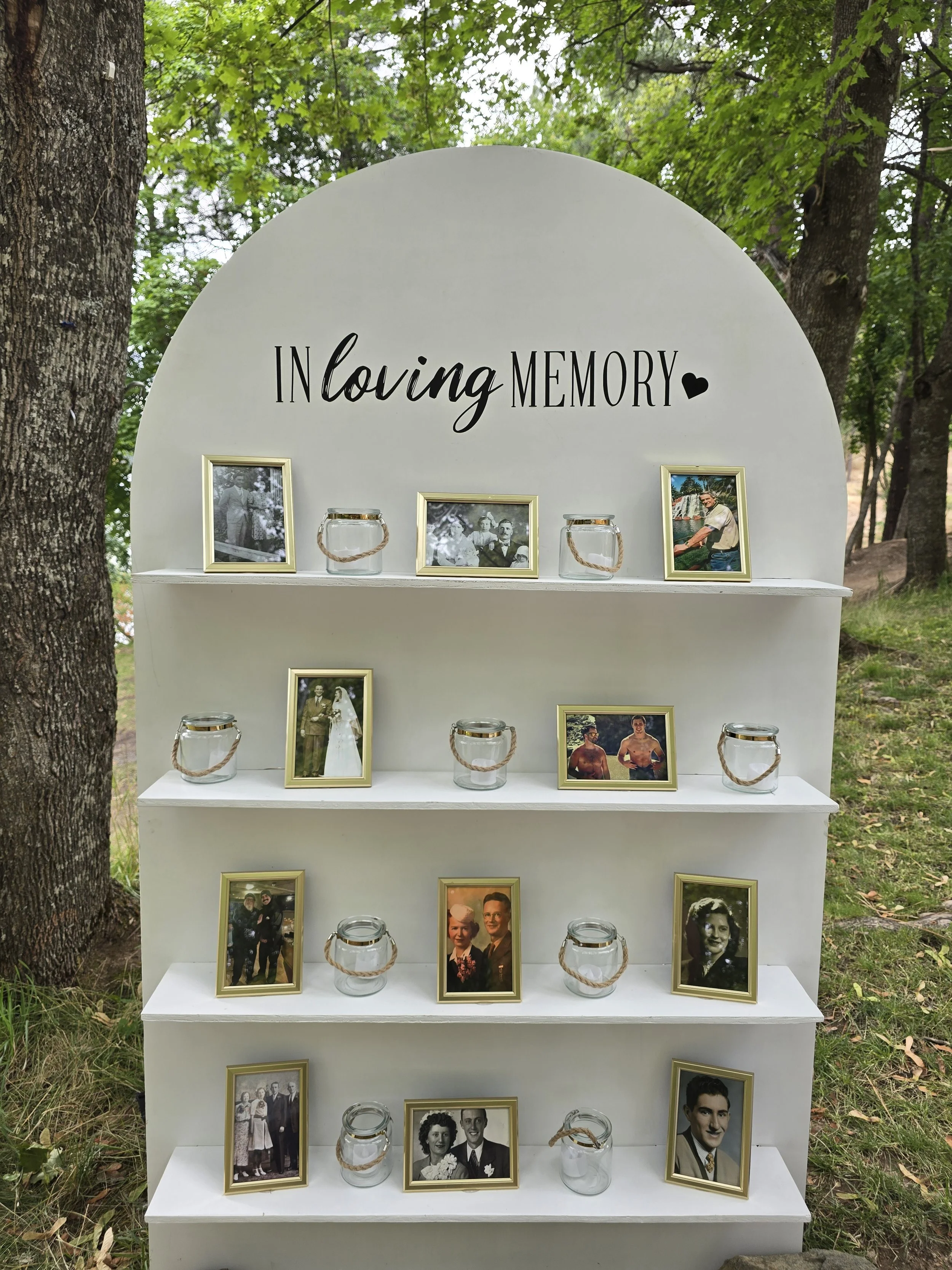 Outdoor memory display titled 'In Loving Memory' with framed photos and small glass candles on white shelves, set among trees.