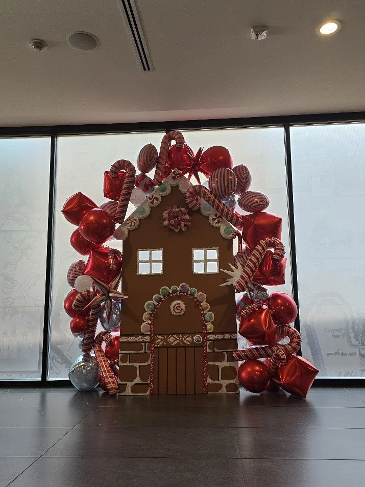 Decorative gingerbread house with red and white candy-themed balloons and ornaments, set against large windows.