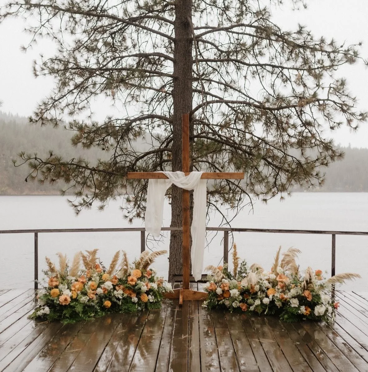 A wooden cross draped with white cloth, set up on a deck by a lake with a tree behind it, surrounded by flower arrangements with peach and white flowers and pampas grass.