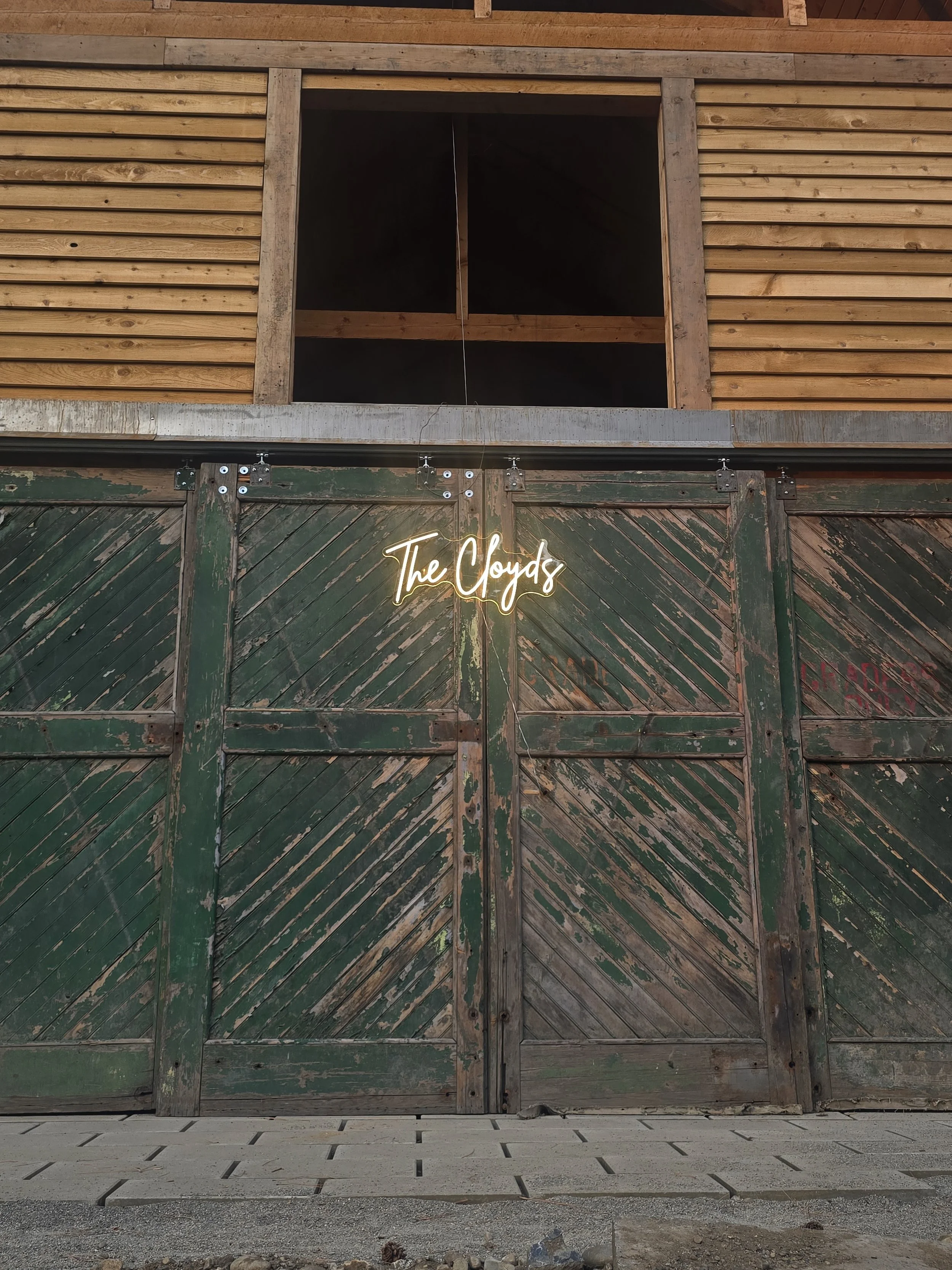 Rustic green barn doors with distressed paint, neon sign saying 'The Cloyds' above the doors, and a wooden upper level with an open window.