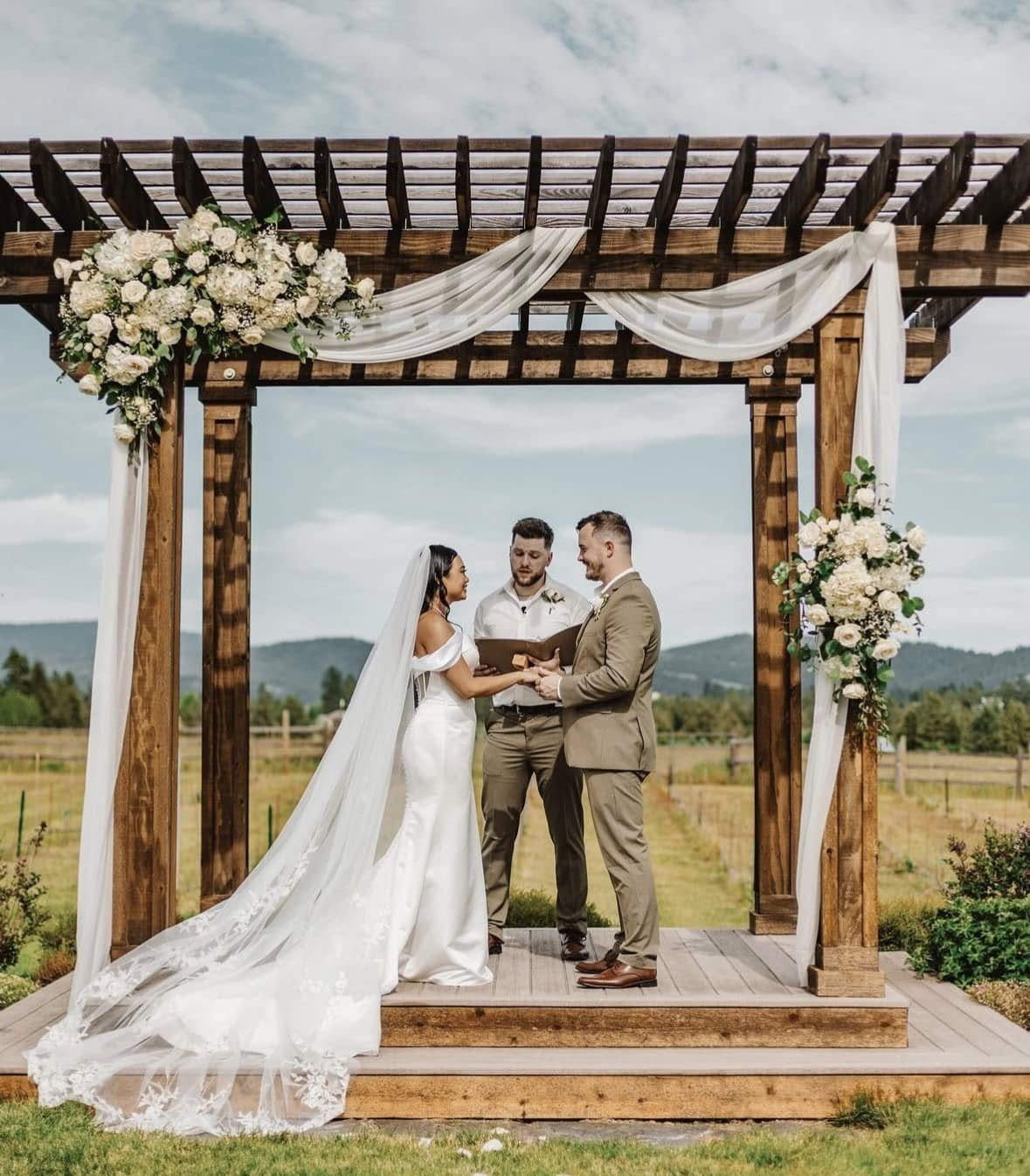 A couple gets married outdoors under a wooden arbor decorated with white flowers and draped white fabric, with mountains and a field in the background.