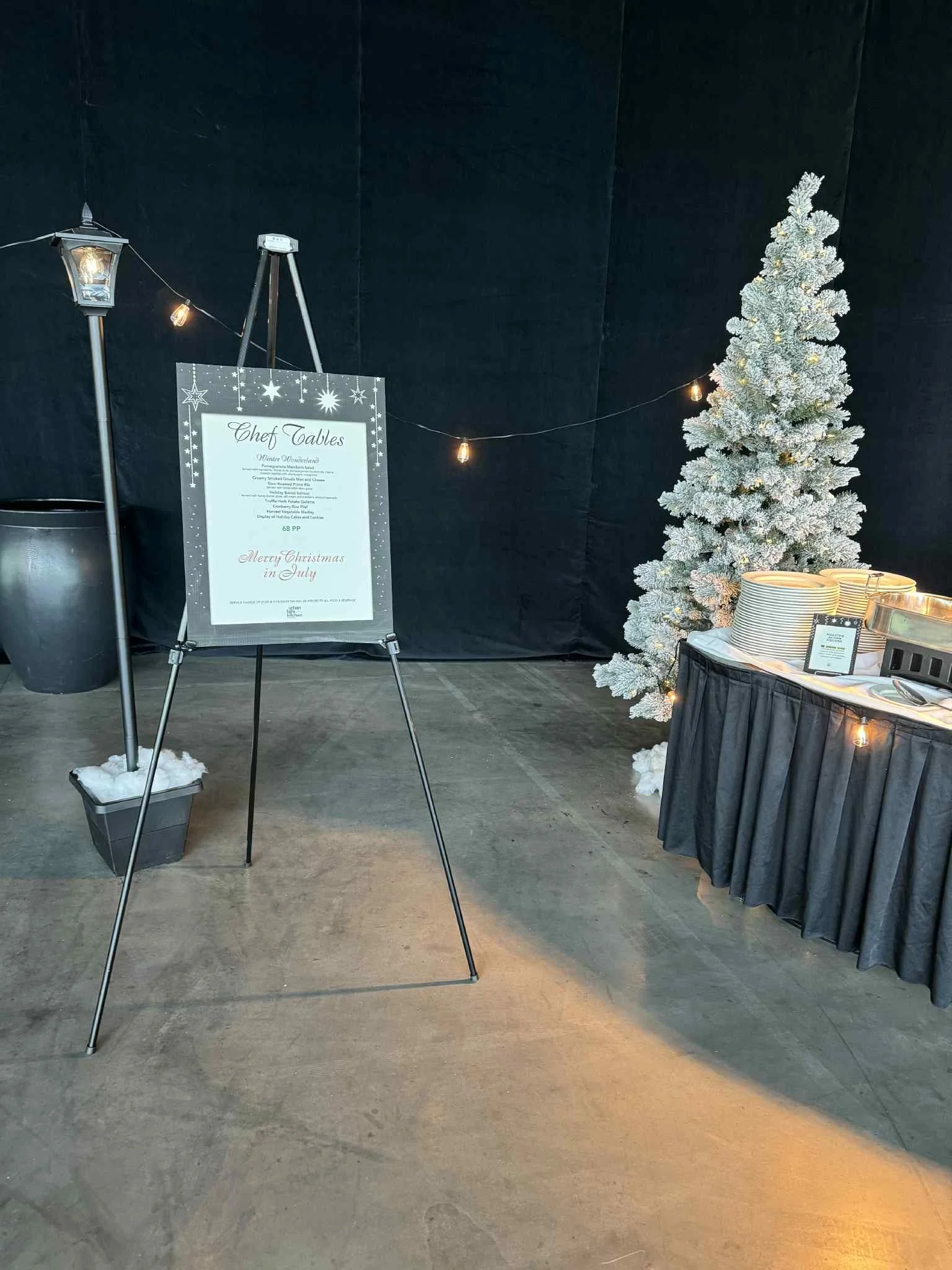 Christmas-themed indoor setup featuring a snow-covered artificial tree, a black table with stacked plates and utensils, and a sign on an easel with a holiday menu, decorated with string lights.