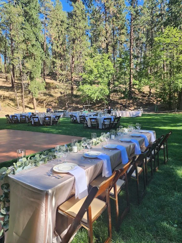 Outdoor wedding reception setup with a long head table decorated with greenery and white cloth, surrounded by chairs, in a grassy area with trees and a wooded background.