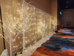 Decorative panel with illuminated white tree branches and fairy lights, with snow-like cotton at the base, in an indoor space.