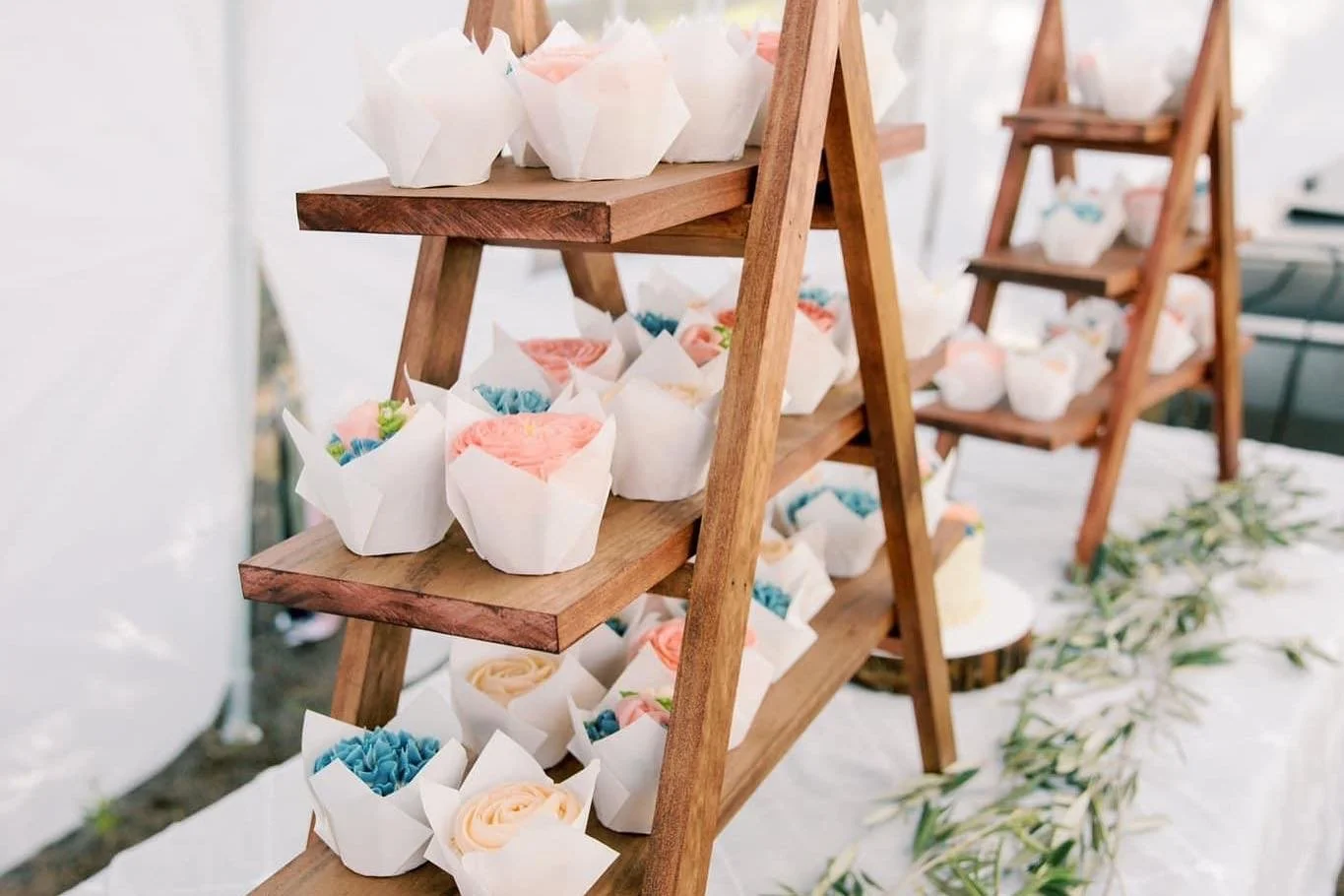 Wooden display shelves holding pastel-colored cupcakes in white paper cups, arranged on a white surface with greenery.